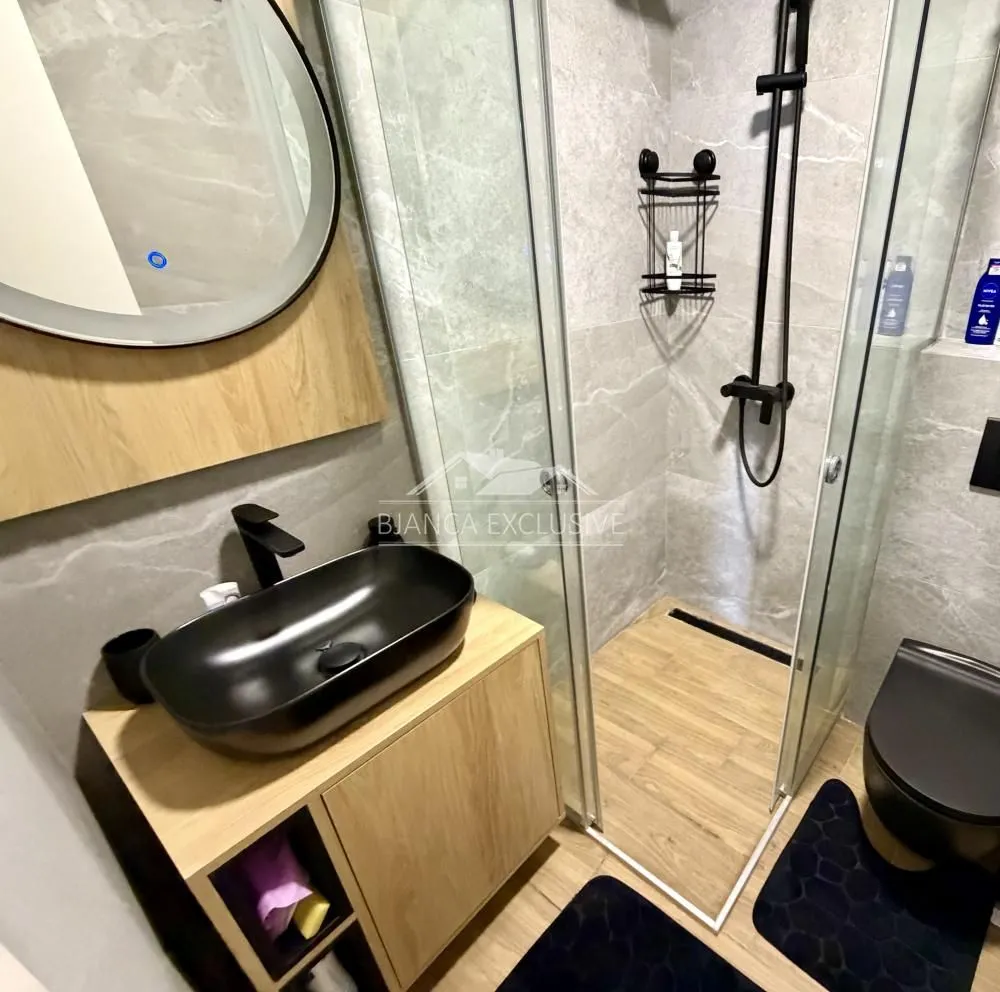 Moderno opremljeno kupatilo sa tuš kabinom, umivaonikom i crnim detaljima.