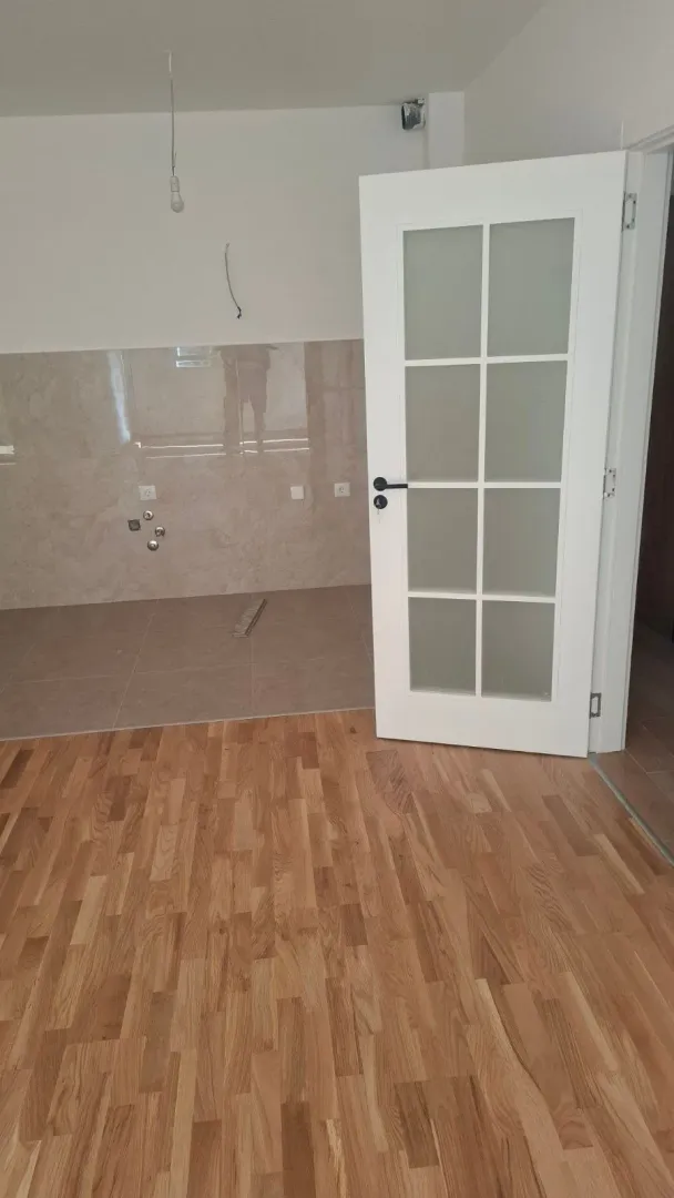 Pogled na dio prostorije sa drvenim podom, keramičkim pločicama na zidu i bijelim vratima.
