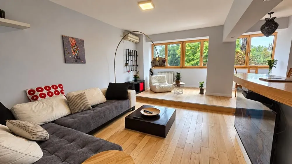 Spacious living room with a sofa set, wooden flooring, and a raised section with windows.