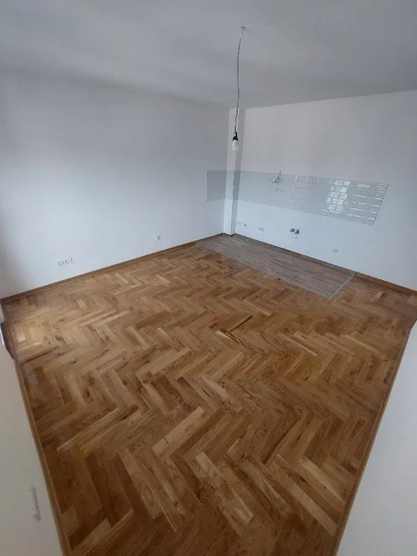 Prazna prostorija sa parketom i pripremljenim delom za kuhinju sa zidnim pločicama.