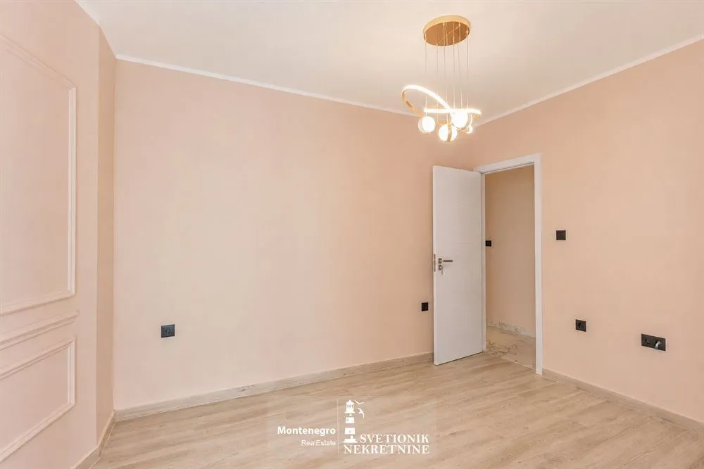 Prazna soba sa svijetlim zidovima, drvenim podom i modernim lusterom.