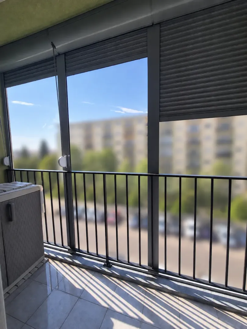 Zastakljeni balkon sa roletnama i pogledom na okolne zgrade i parking.
