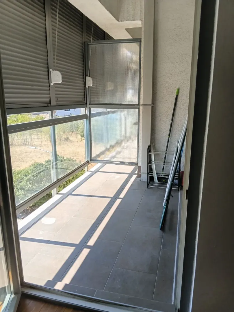 Zastakljeni balkon sa keramičkim pločicama na podu i pogledom na zelenilo.