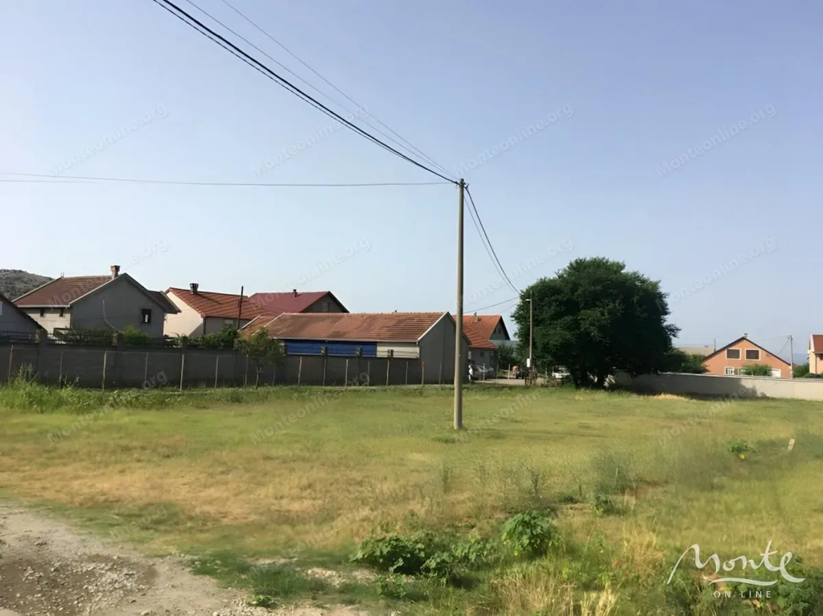 Pogled na plac sa travnatom površinom i kućama u pozadini.