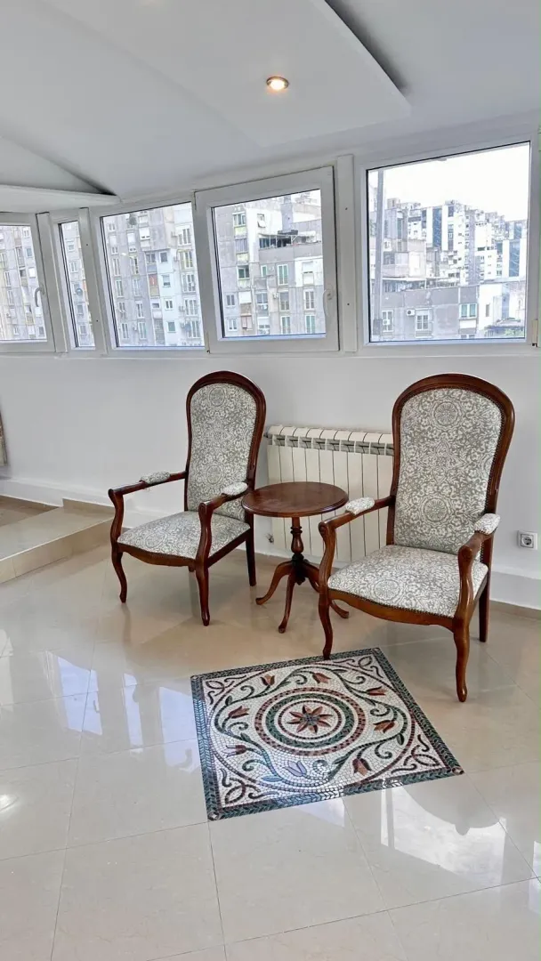 A corner arranged with two armchairs and a small table by large windows with a city view.
