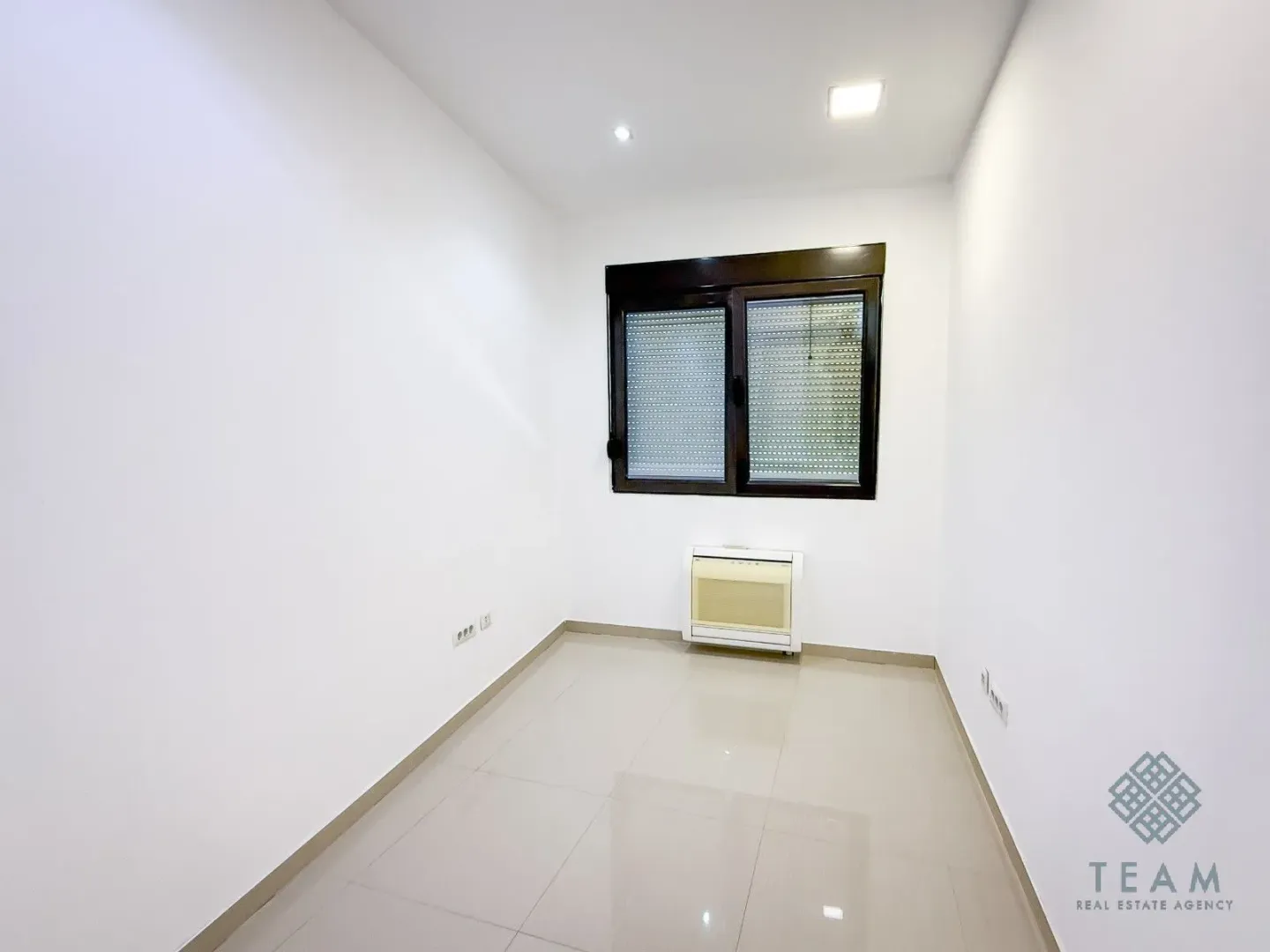 Empty room with white walls, tiled floor, and a window with roller shutters.