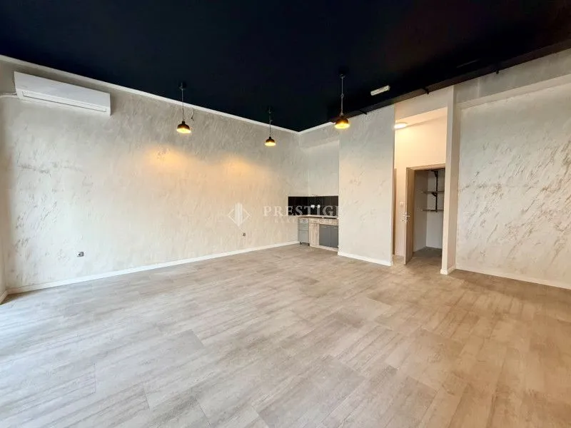 Spacious empty room with light walls and a kitchen in the background.