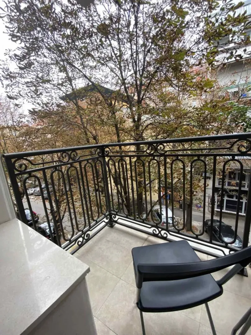 Small furnished balcony with wrought iron railing and a view of trees and the street.