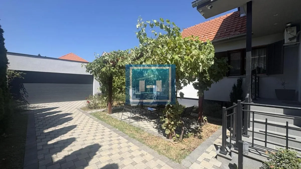 Dvorište kuće sa popločanim prilazom, garažom i sjenicom prekrivenom lozom.