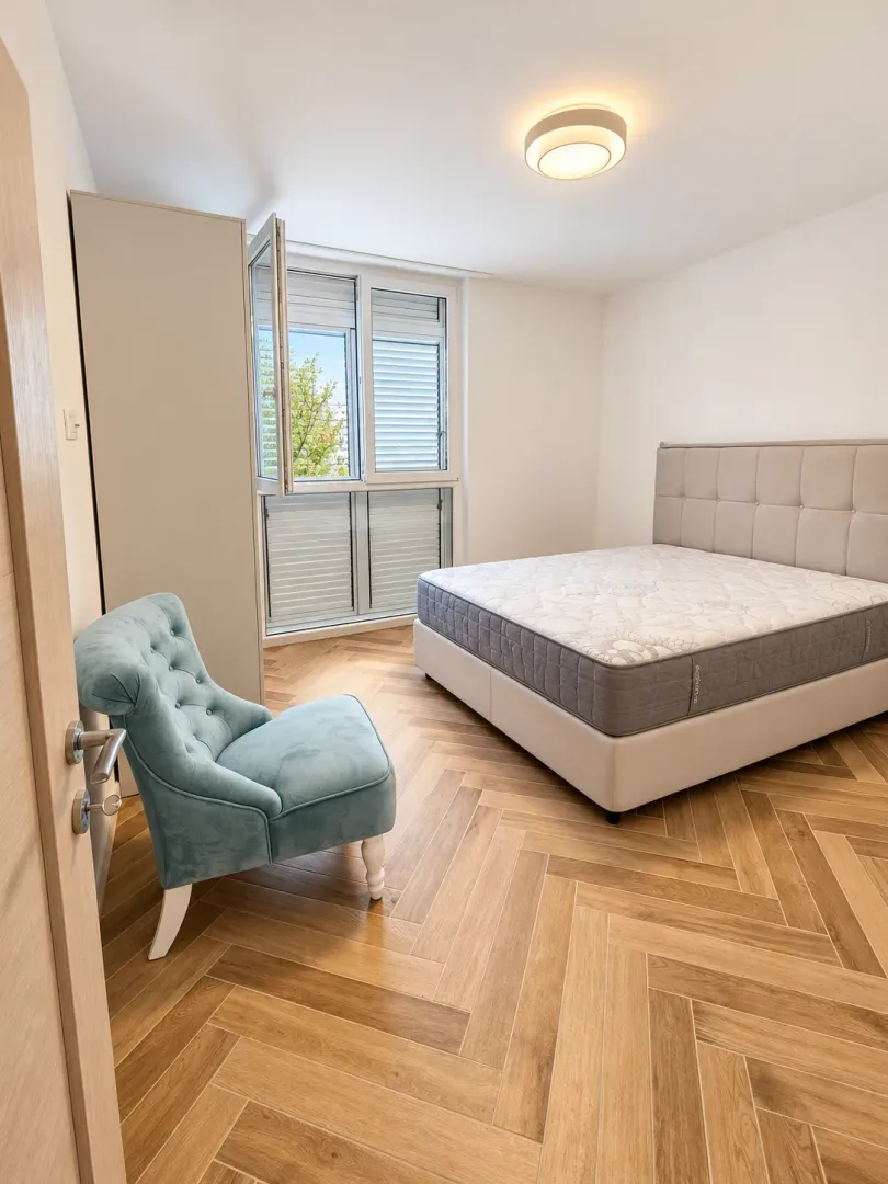 Spavaća soba sa bračnim krevetom, plavom foteljom i parketom u riblju kost.