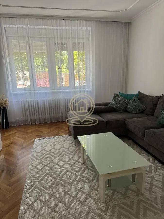 Dnevna soba sa ugaonom garniturom, stočićem i prozorom sa zavesama.