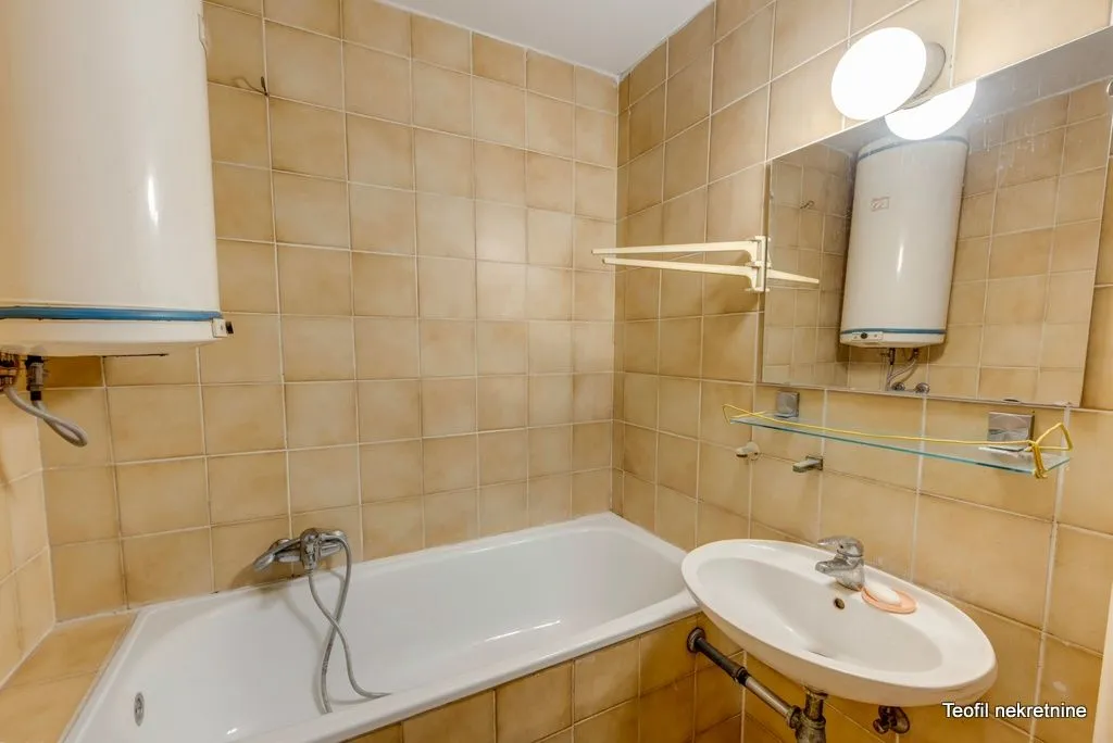 Kupatilo sa kadom, umivaonikom, ogledalom i bojlerom obloženo bež pločicama.