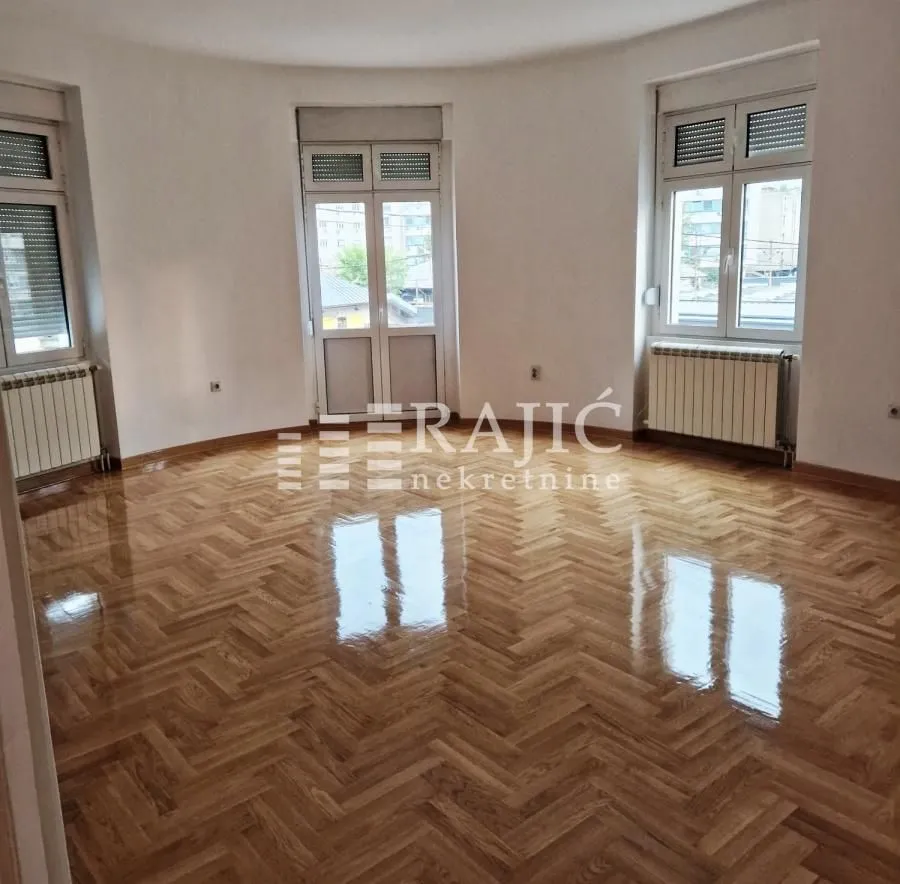 Empty room with parquet flooring and large windows.