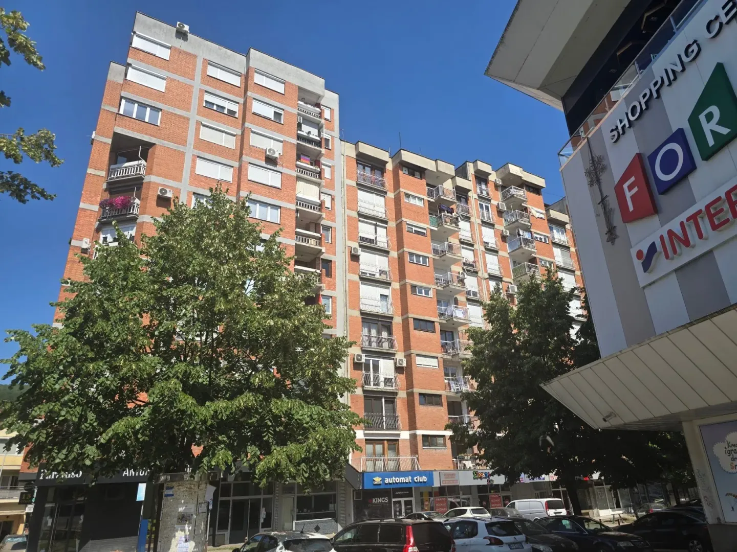 Spoljašnji izgled stambene zgrade sa okolnim drvećem i parkingom.