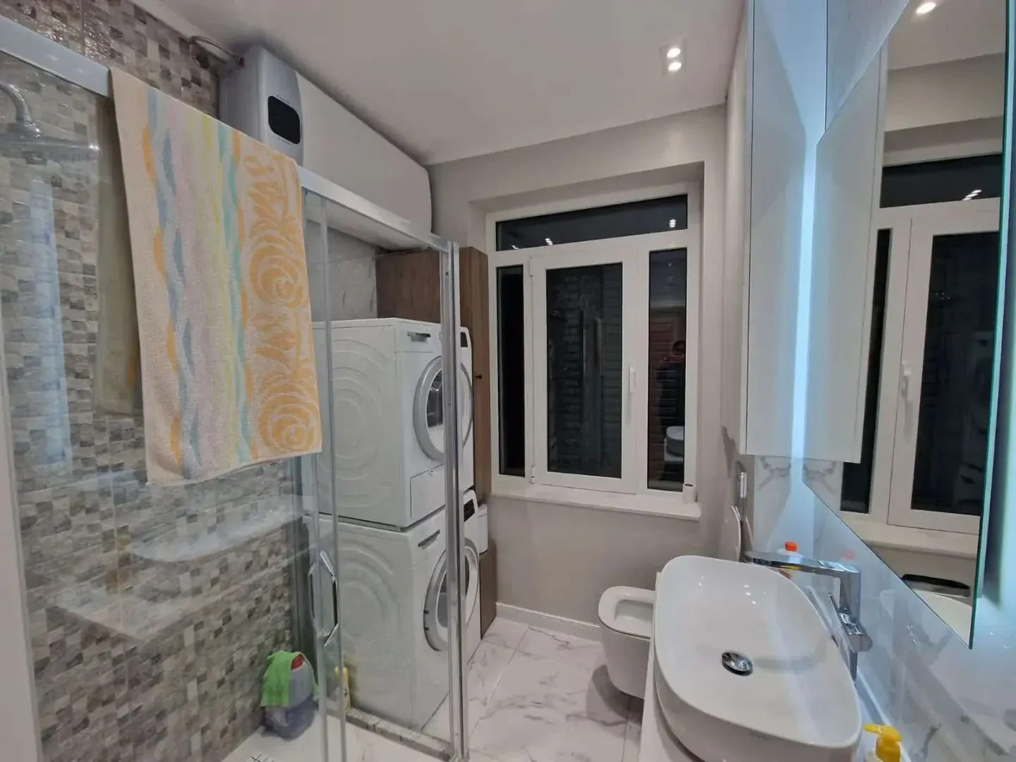 Modern bathroom with a walk-in shower, washbasin, and stacked washing machine.