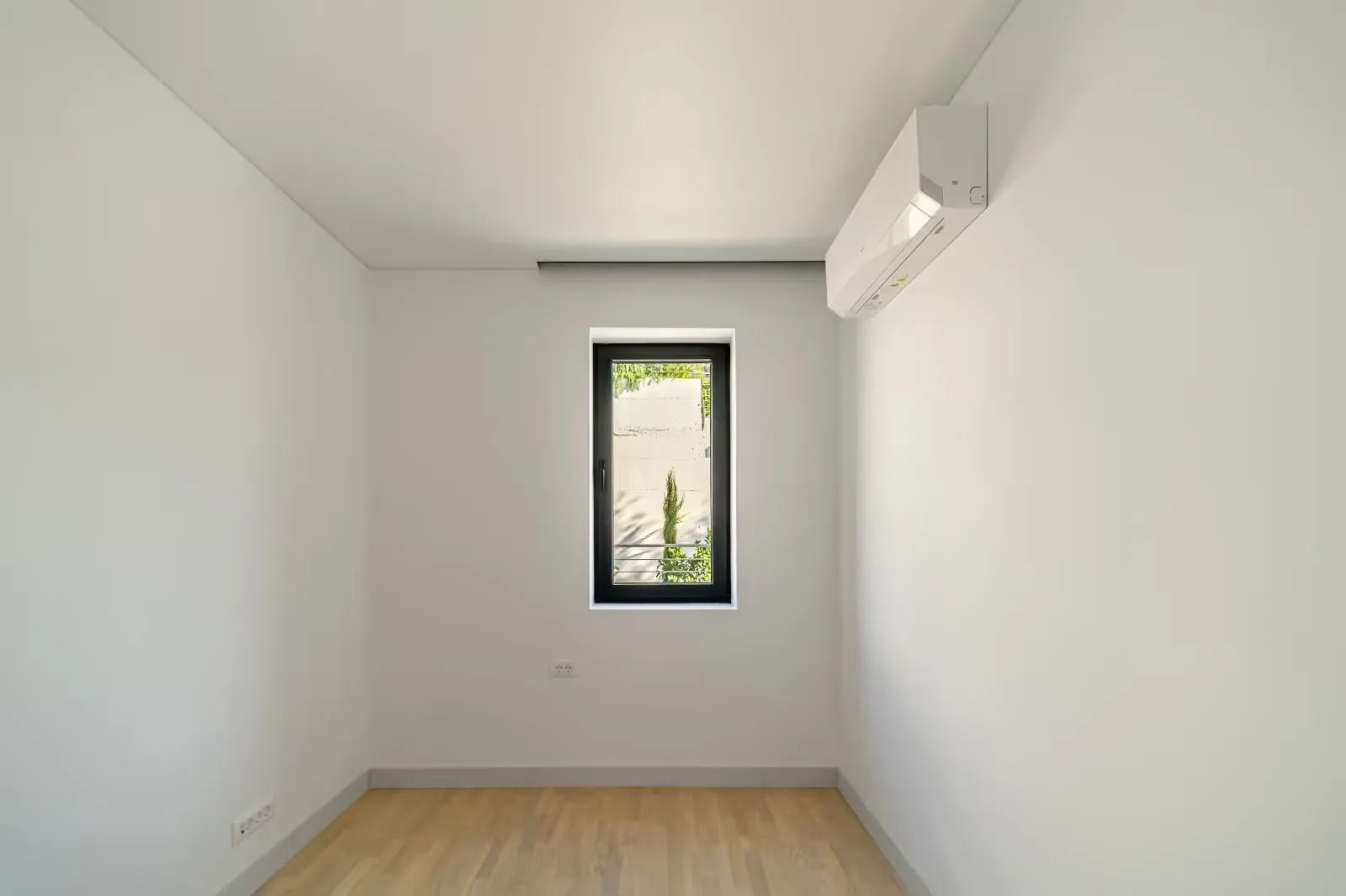 Prazna soba sa bijelim zidovima, drvenim podom, prozorom i klima uređajem.