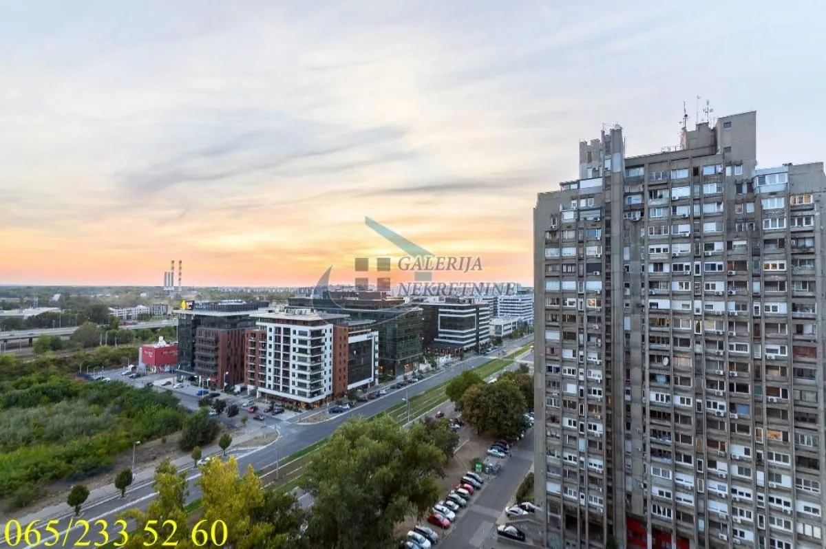 Pogled sa prozora na gradski pejzaž i okolne zgrade tokom zalaska sunca.
