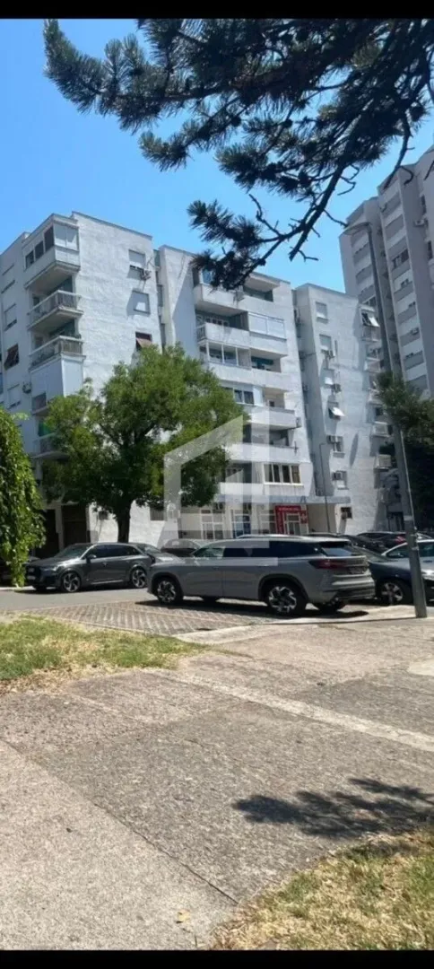 Pogled na spoljašnost stambene zgrade i parking sa vozilima.