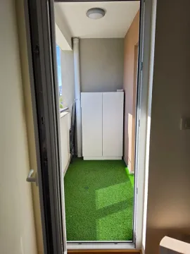 Pogled kroz balkonska vrata na terasu sa vještačkom travom i ormarom. - image 13