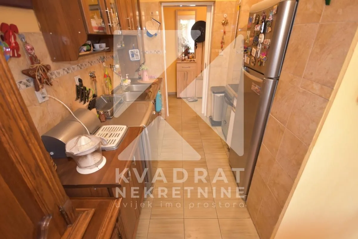 Pogled na usku kuhinju sa elementima i frižiderom.