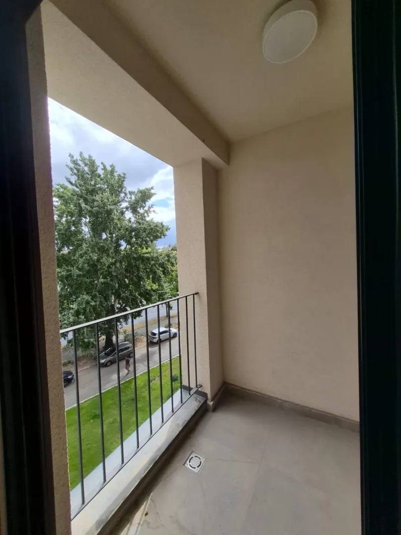 Prazan natkriveni balkon sa metalnom ogradom i pogledom na ulicu i drveće.