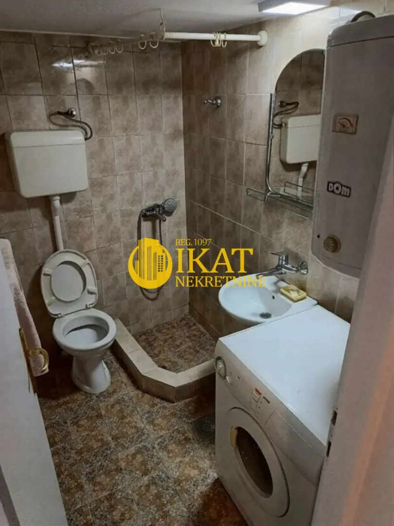 Manje kupatilo obloženo keramičkim pločicama sa tuš kabinom, WC šoljom, umivaonikom i mašinom za veš.