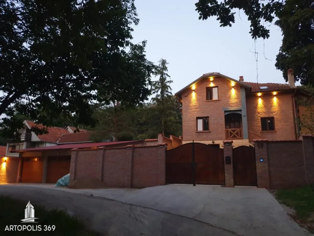 Pogled na spoljašnjost kuće sa dvorištem, kapijom i osvetljenom fasadom u sumrak.
