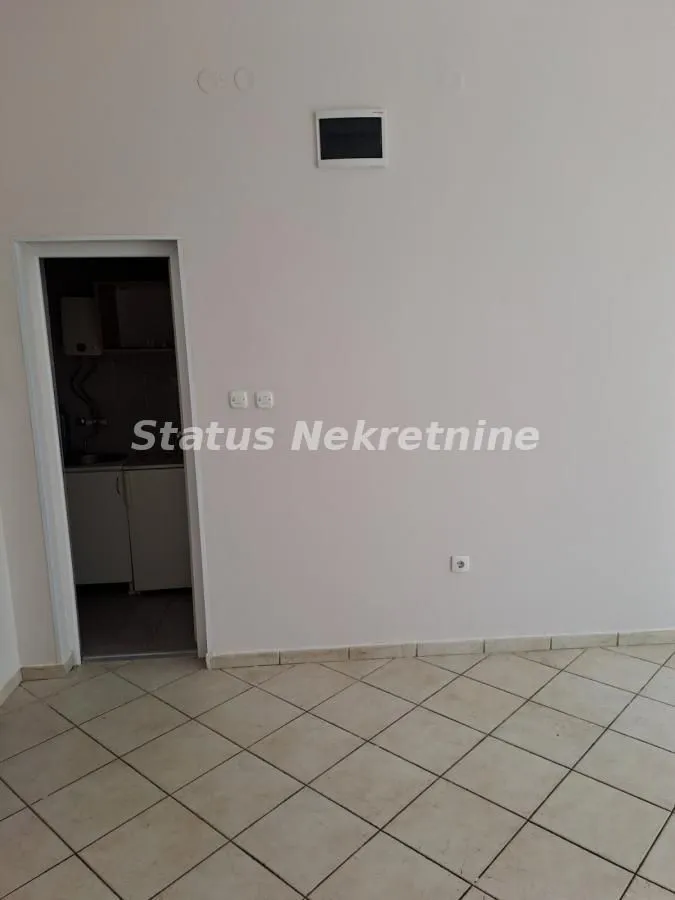 Empty room with ceramic tiles and a view of a small kitchen through the doorway.