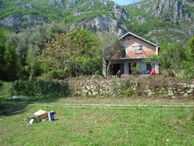 Nedovršena kuća okružena zelenilom i drvećem u podnožju brda.