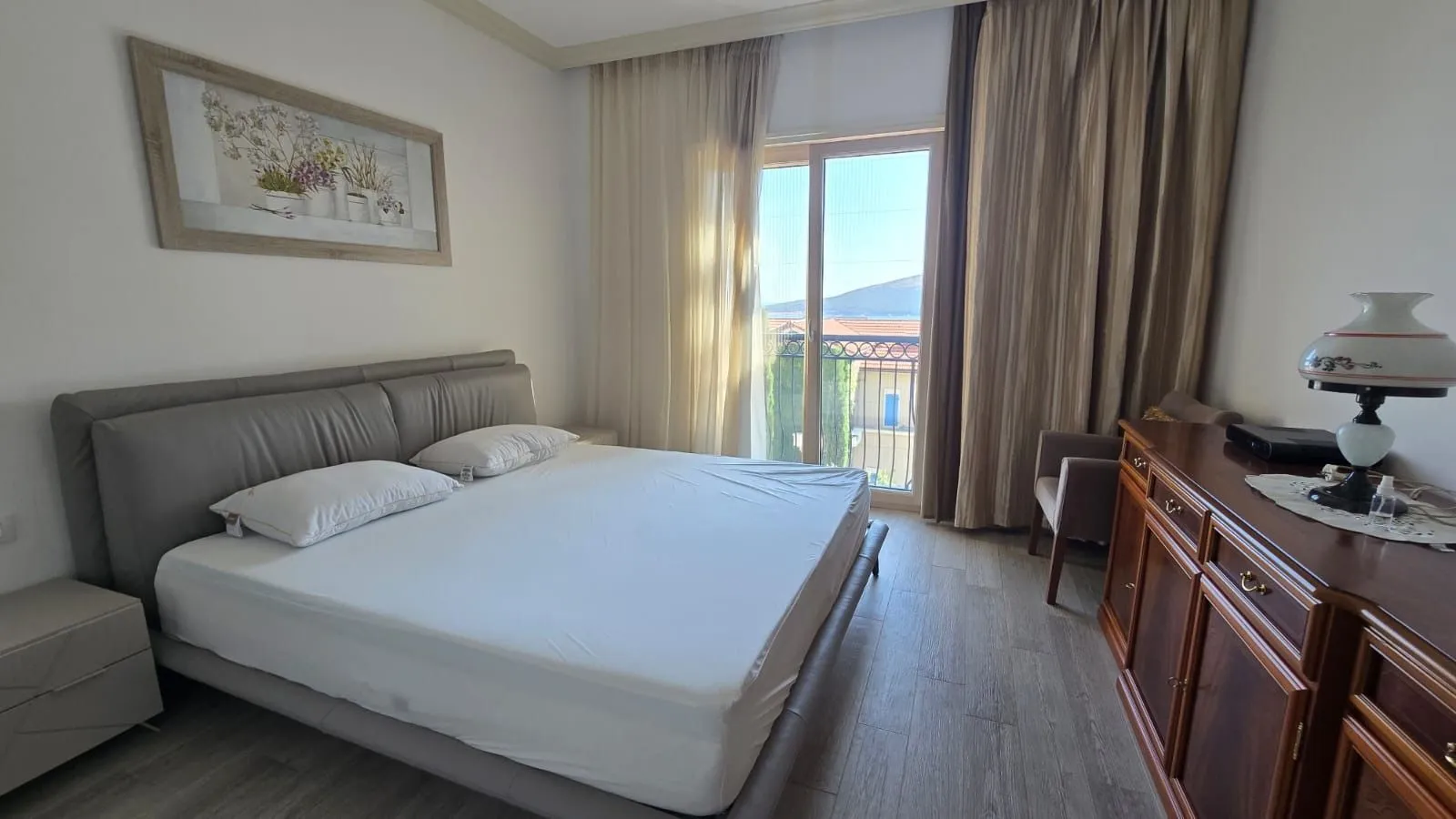Spavaća soba sa bračnim krevetom, drvenom komodom i izlazom na balkon.