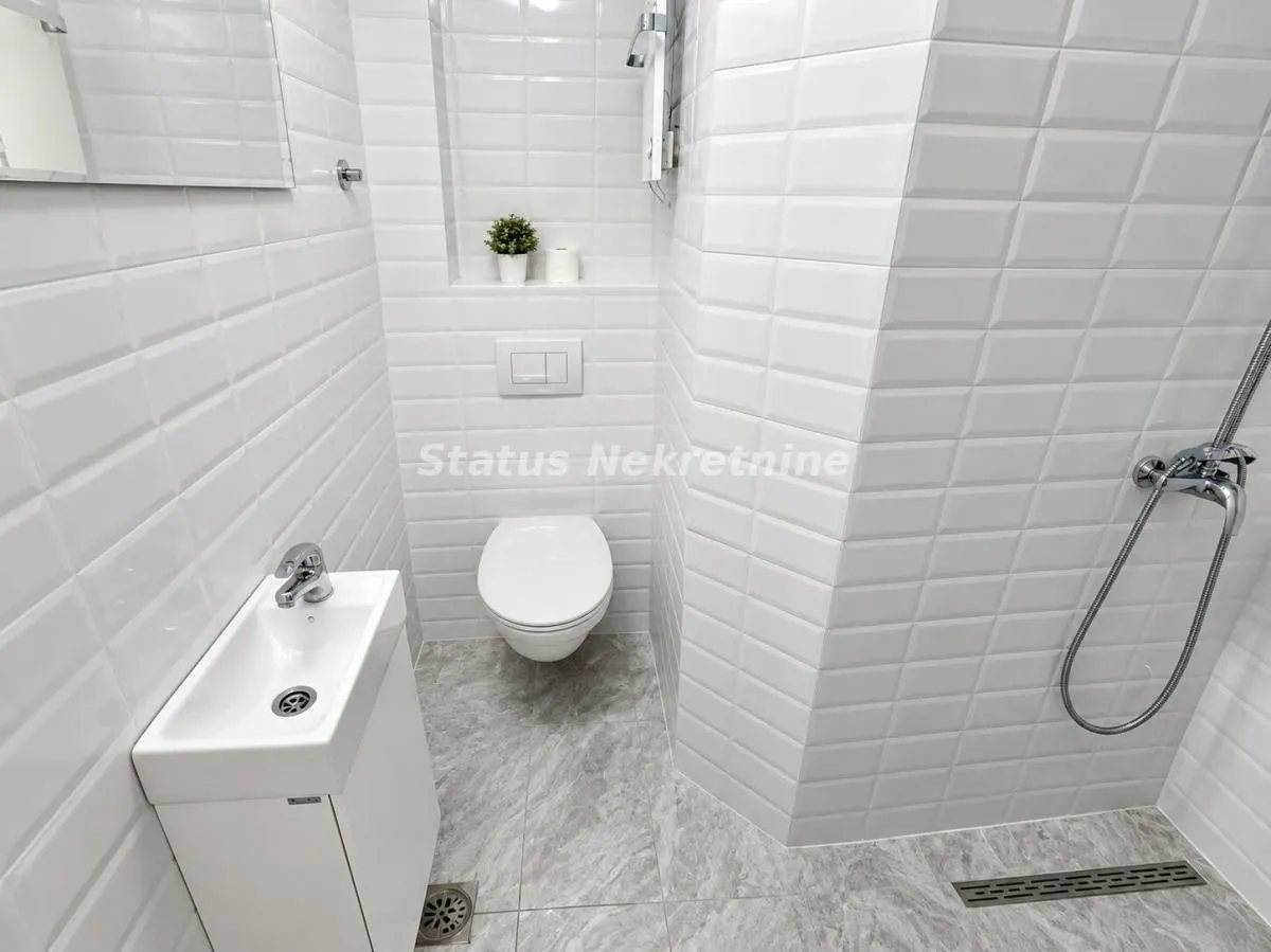 Manje kupatilo obloženo belim keramičkim pločicama sa tuš kabinom.
