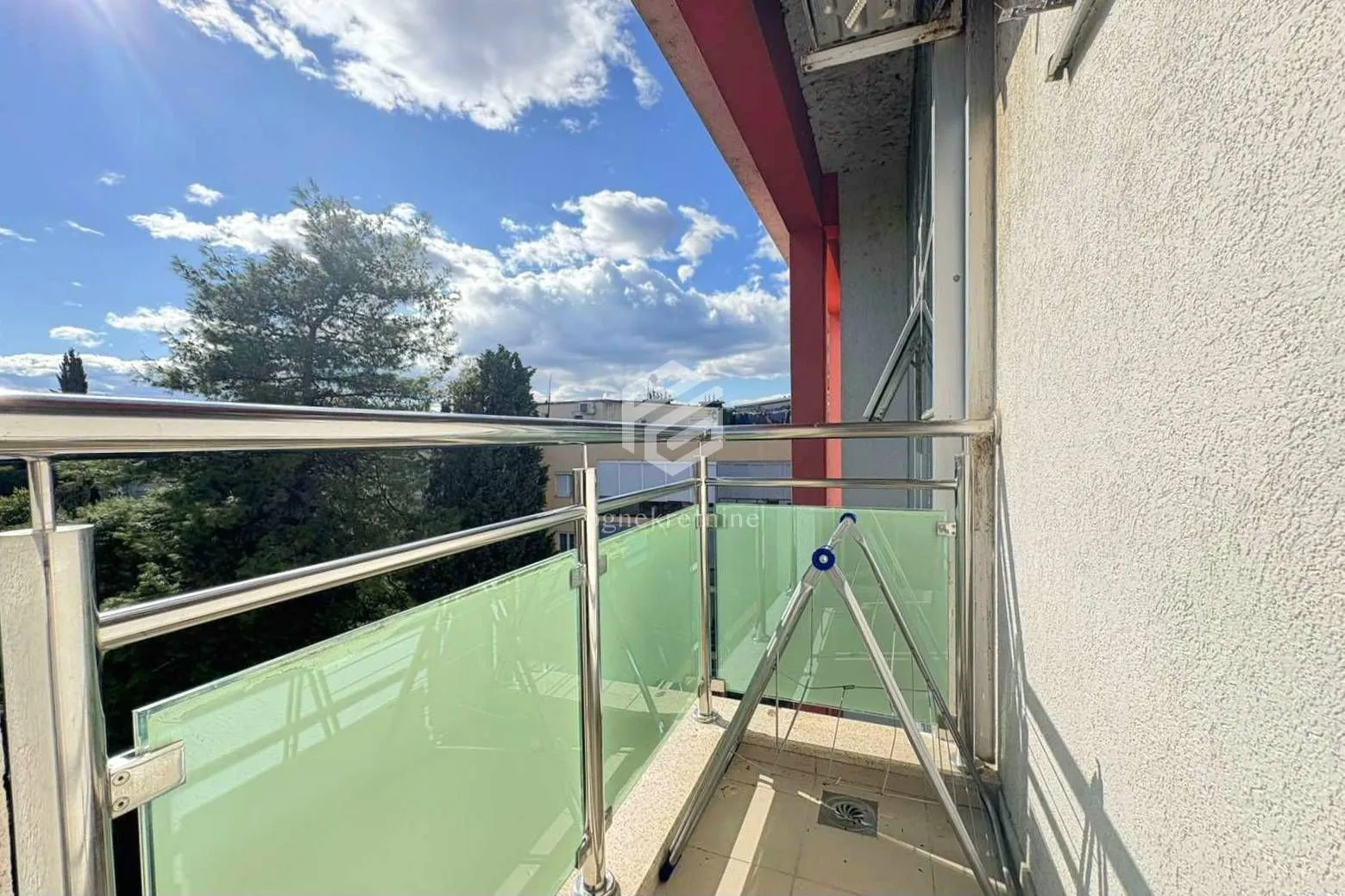 Manji balkon sa staklenom ogradom i pogledom na zelenilo.