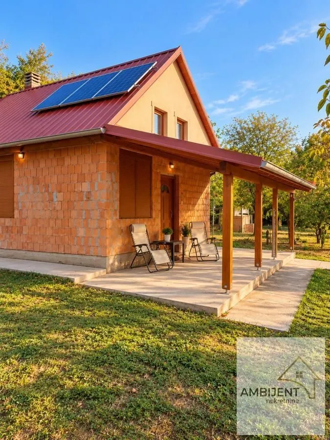 Pogled na spoljašnjost kuće sa natkrivenom terasom, dvorištem i solarnim panelima na krovu.