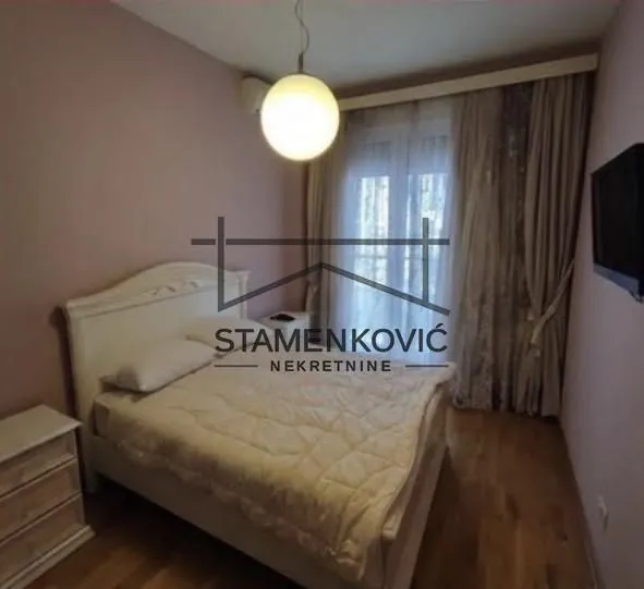 Nameštena spavaća soba sa bračnim krevetom, noćnim stočićem i prozorom sa zavesama.