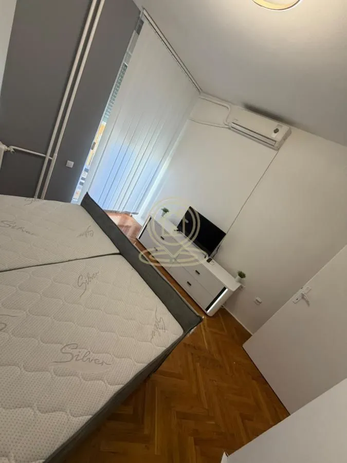 Nameštena spavaća soba sa bračnim krevetom, komodom za televizor i klima uređajem.