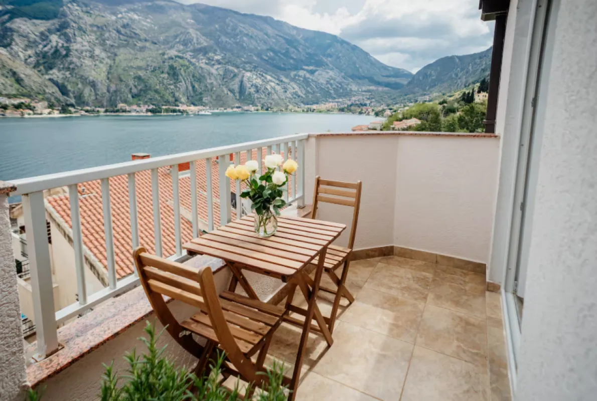Balkon sa drvenim stolom, stolicama i vazom sa cvijećem, sa pogledom na more i planine.