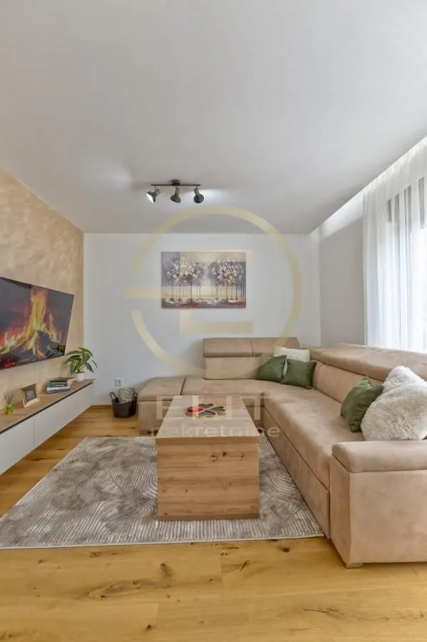 Dnevna soba sa ugaonom garniturom, televizorom i drvenim podom.