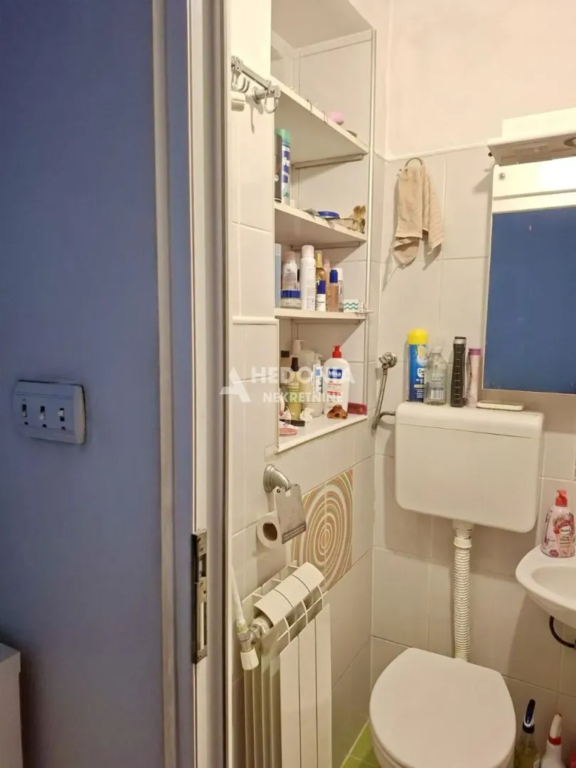 Manje kupatilo sa wc šoljom, umivaonikom, ogledalom i ugradnim policama na zidu.