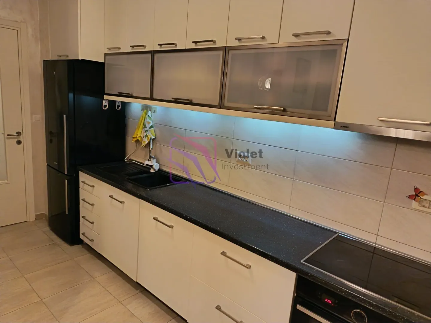 Kuhinja sa bijelim elementima, tamnom radnom pločom i crnim frižiderom.