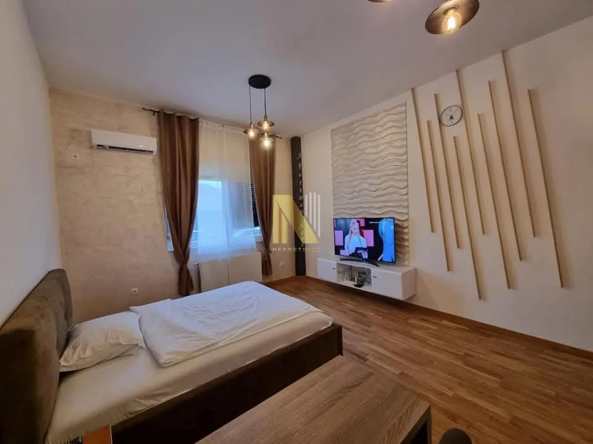Nameštena spavaća soba sa bračnim krevetom, televizorom i dekorativnim drvenim zidnim panelima.