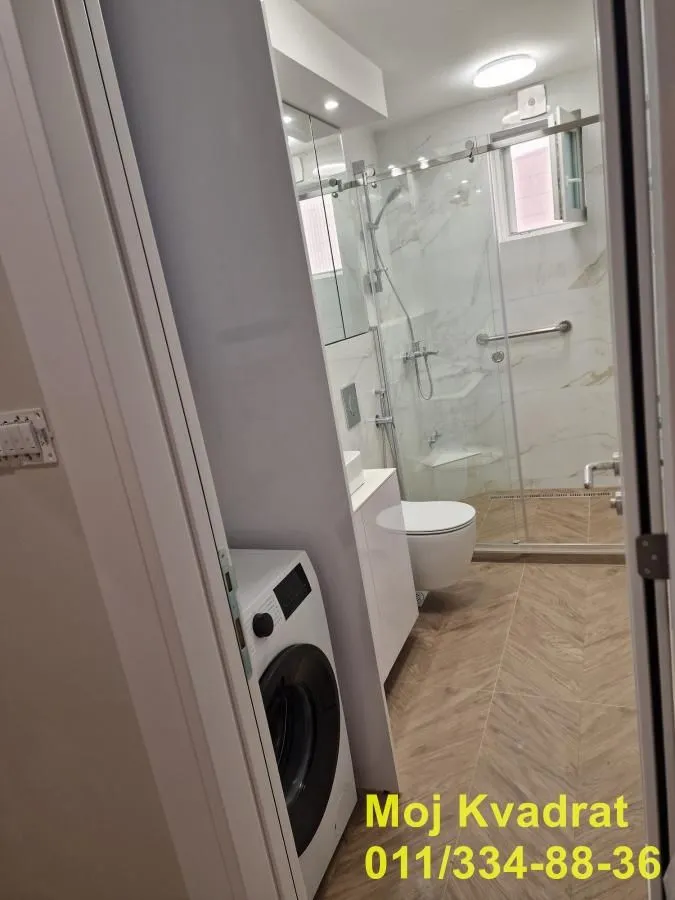 Pogled iz hodnika na moderno opremljeno kupatilo sa tuš kabinom i veš mašinom.