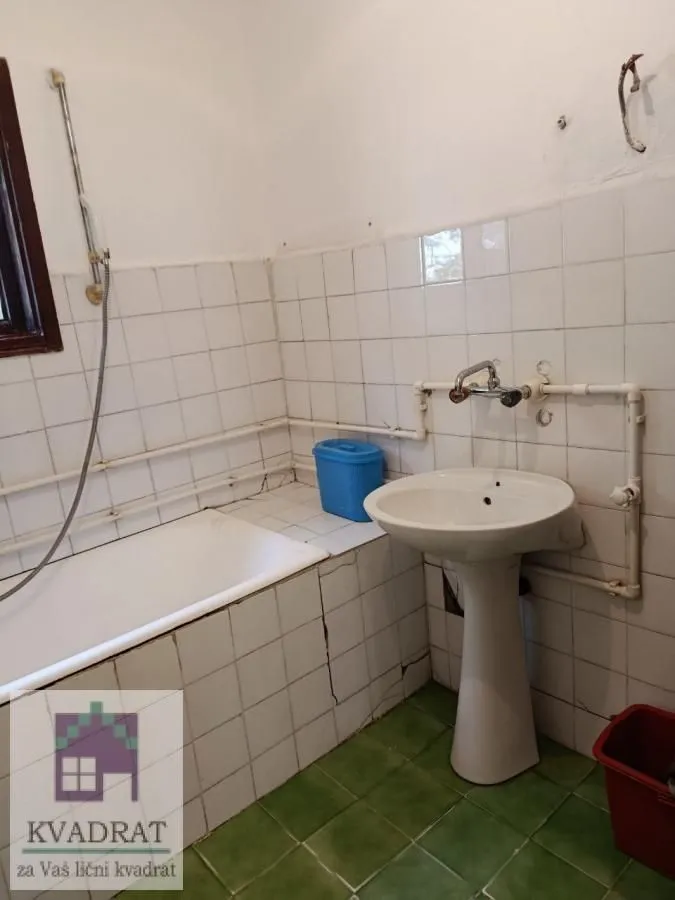Starije opremljeno kupatilo sa kadom i umivaonikom obloženo belim pločicama.