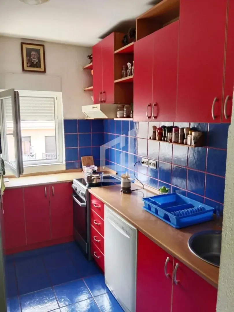Kuhinja sa crvenim elementima, plavim pločicama i prozorom.
