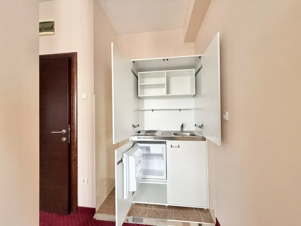 Small built-in kitchen unit with open doors and a refrigerator in the hallway.