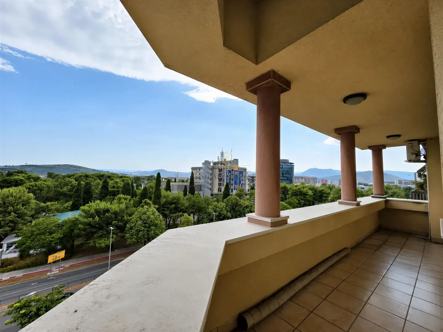 Spacious covered balcony with columns and a view of urban greenery and surroundings.