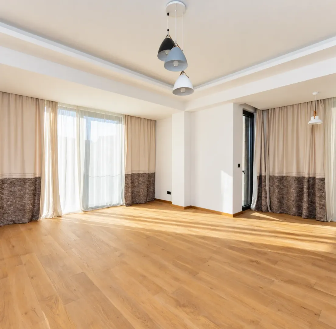 Empty living room with hardwood floors, large windows, and modern lighting.