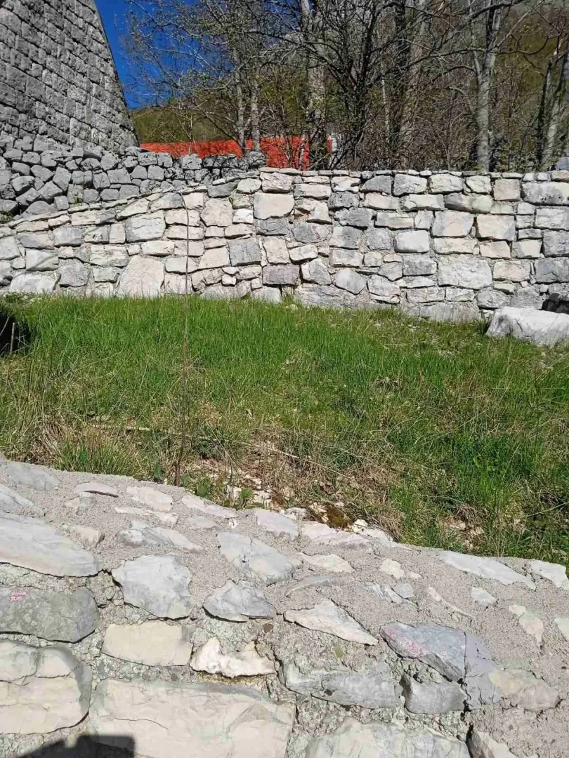 Kameni potporni zidovi i travnati dio placa sa drvećem u pozadini.