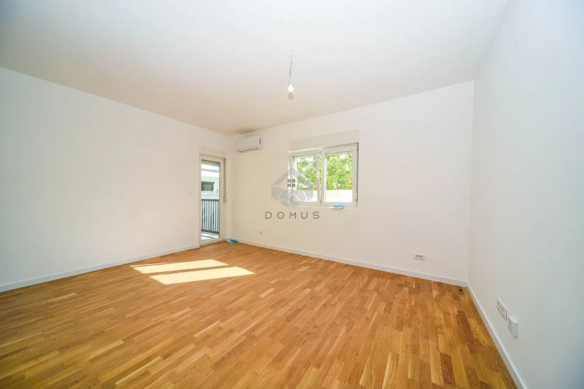 Prazna soba sa bijelim zidovima, drvenim parketom, prozorom i vratima koja vode na terasu.