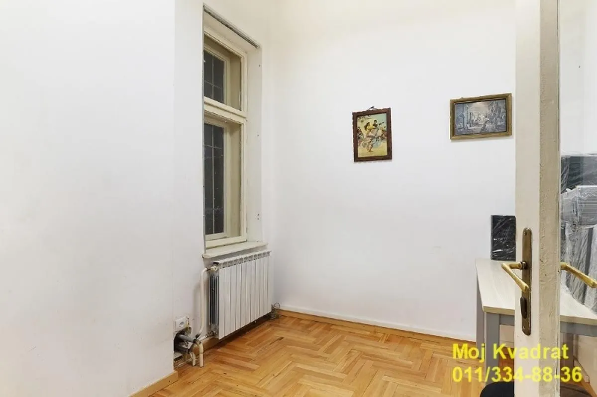 Manja soba sa belim zidovima, parketom, prozorom i radijatorom.