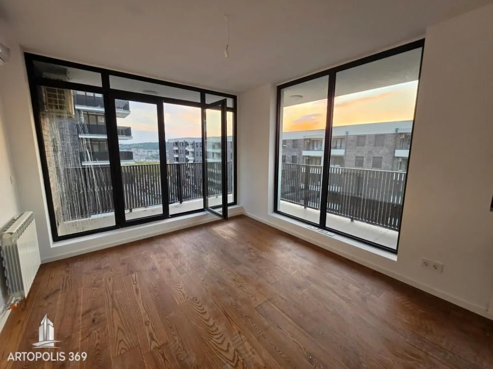 Empty room with wooden flooring and large windows overlooking surrounding buildings.